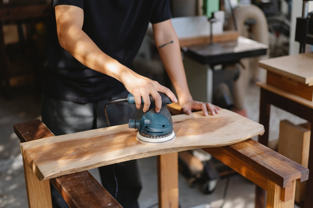 Joinery Services Staffordshire. A craftsman uses a power sander on a wooden plank in a well-lit workshop.
